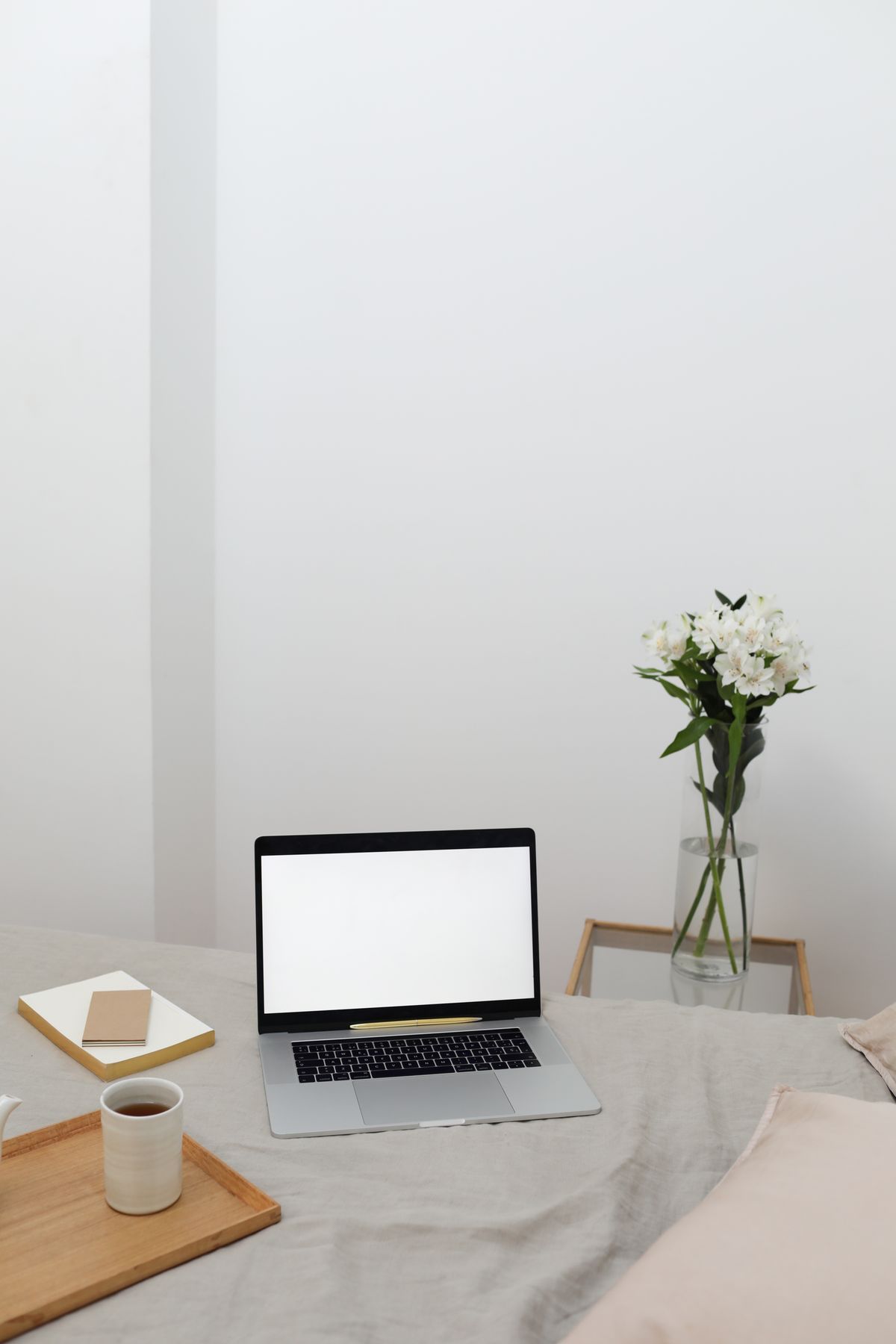 Modern laptop with blank screen and notebooks arranged on soft bed with tray with cup of coffee near table with bunch of fresh flowers during remote work at home