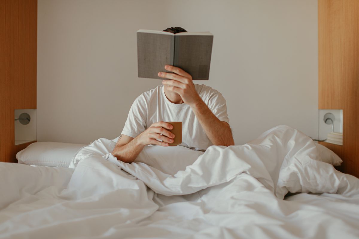 Adult man relaxing and reading a book in bed, holding a cup, cozy morning indoors.