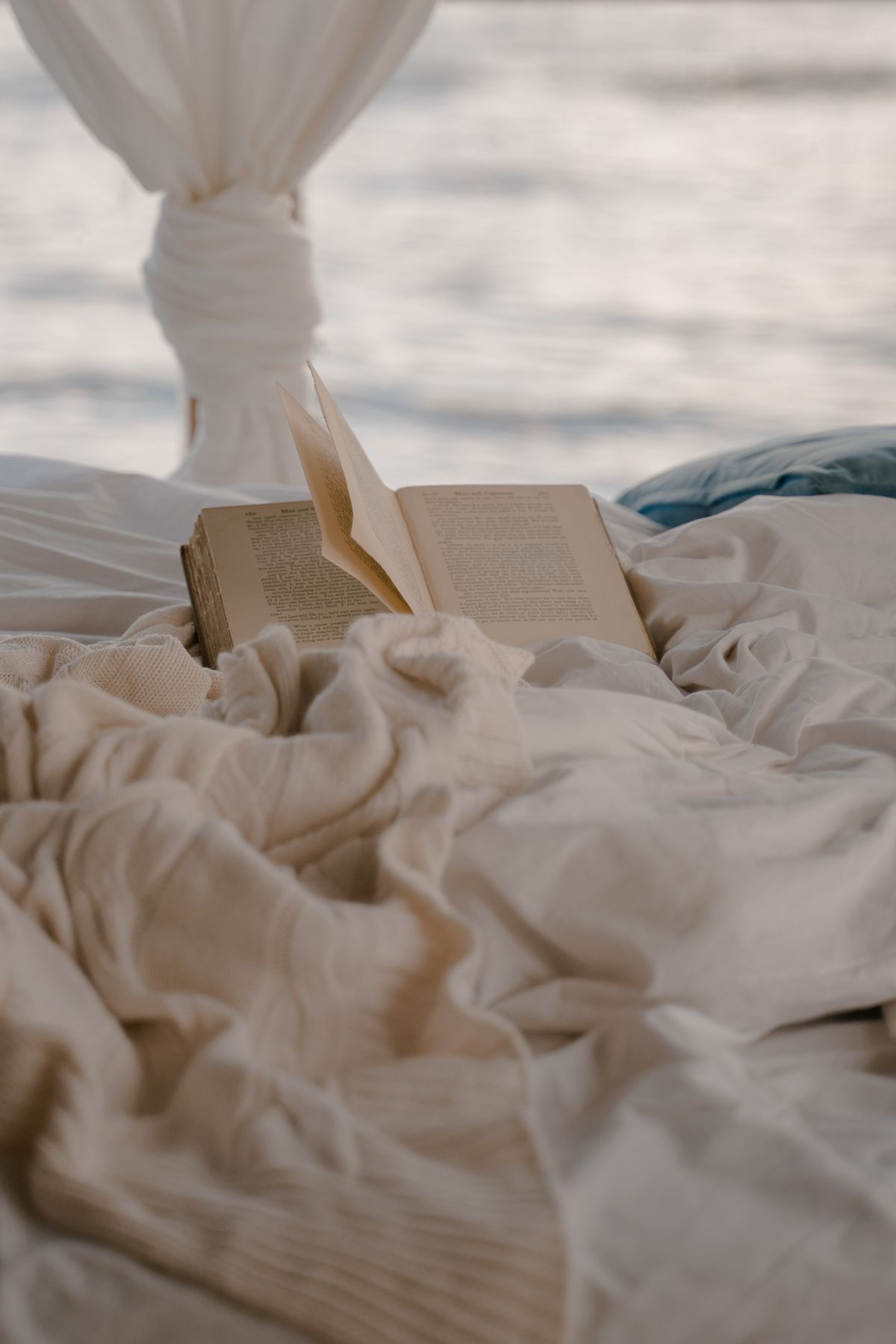Open book resting on a cozy unmade bed with soft lighting and a view through sheer curtains.