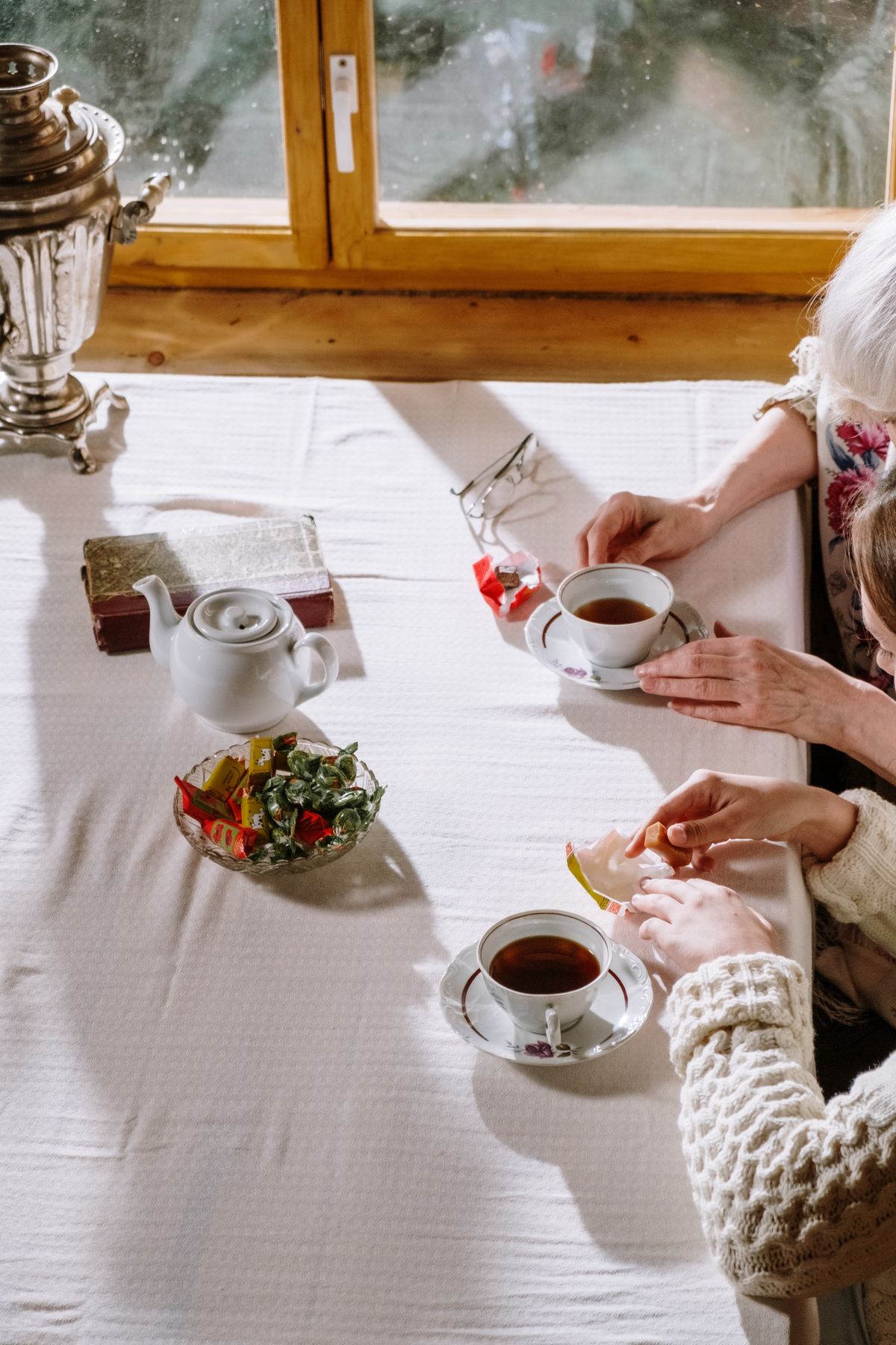 A warm, cozy setting featuring tea cups, sweets, and hands in soft sunlight.